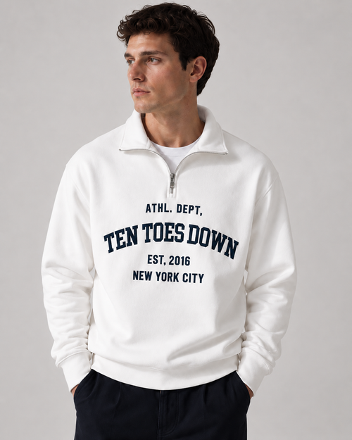 A man wearing a white quarter-zip sweatshirt with navy blue collegiate-style oversized  text reading 'TEN TOES DOWN'