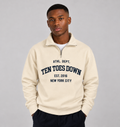 A man wearing a cream-colored quarter-zip sweatshirt with navy blue collegiate-style oversized text reading " TEN TOES DOWN"