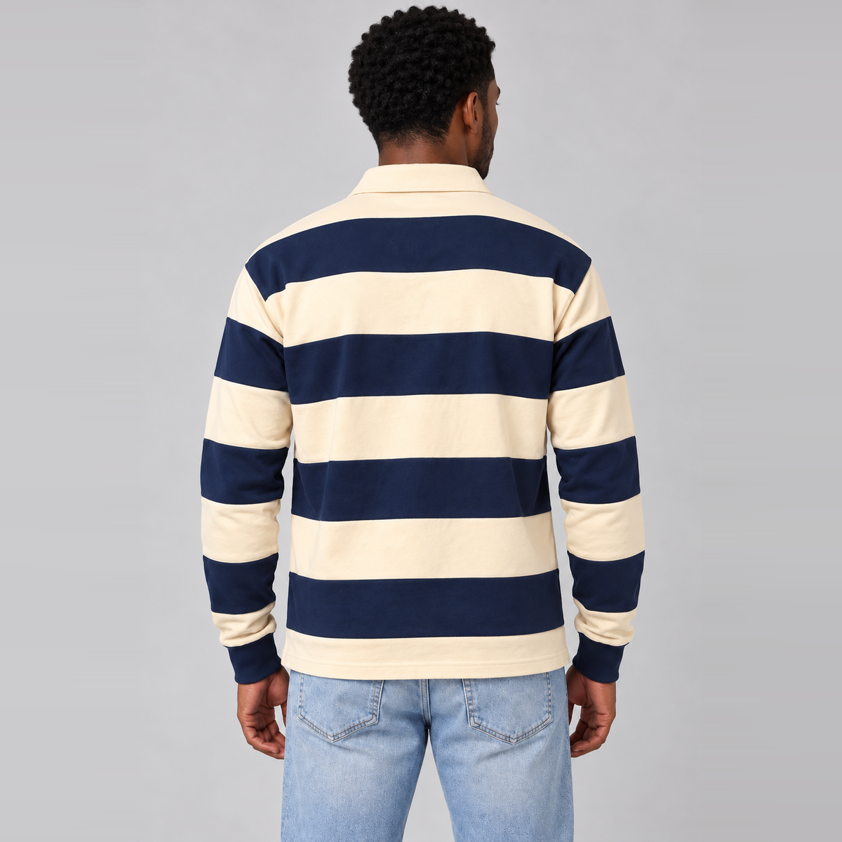 A man viewed from behind wearing a navy blue and cream horizontal striped long-sleeve rugby shirt with light blue jeans, set against a solid gray studio background.