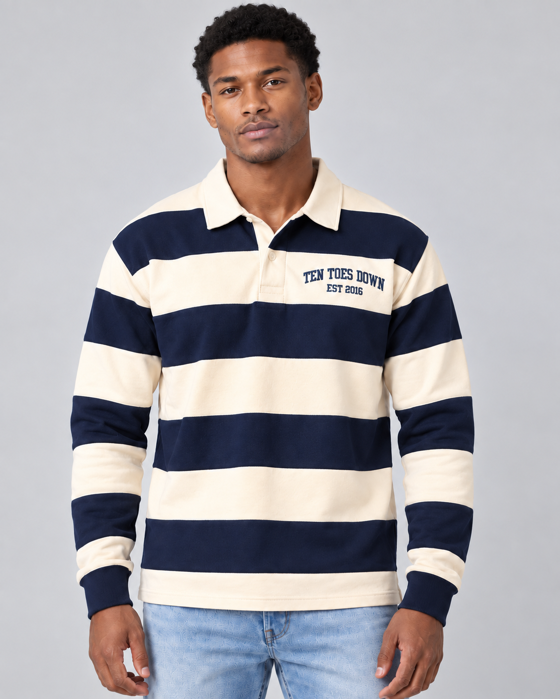 A man wearing a navy blue and cream horizontally striped long-sleeve rugby shirt with 'TEN TOES DOWN EST. 2026' printed on the left chest, paired with light blue jeans, against a solid gray studio background.