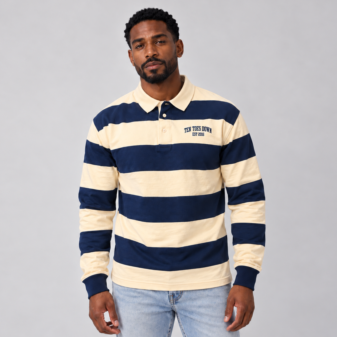 A man wearing a navy blue and cream horizontally striped long-sleeve rugby shirt with 'TEN TOES DOWN EST. 2026' printed on the left chest, paired with light blue jeans, against a solid gray studio background.