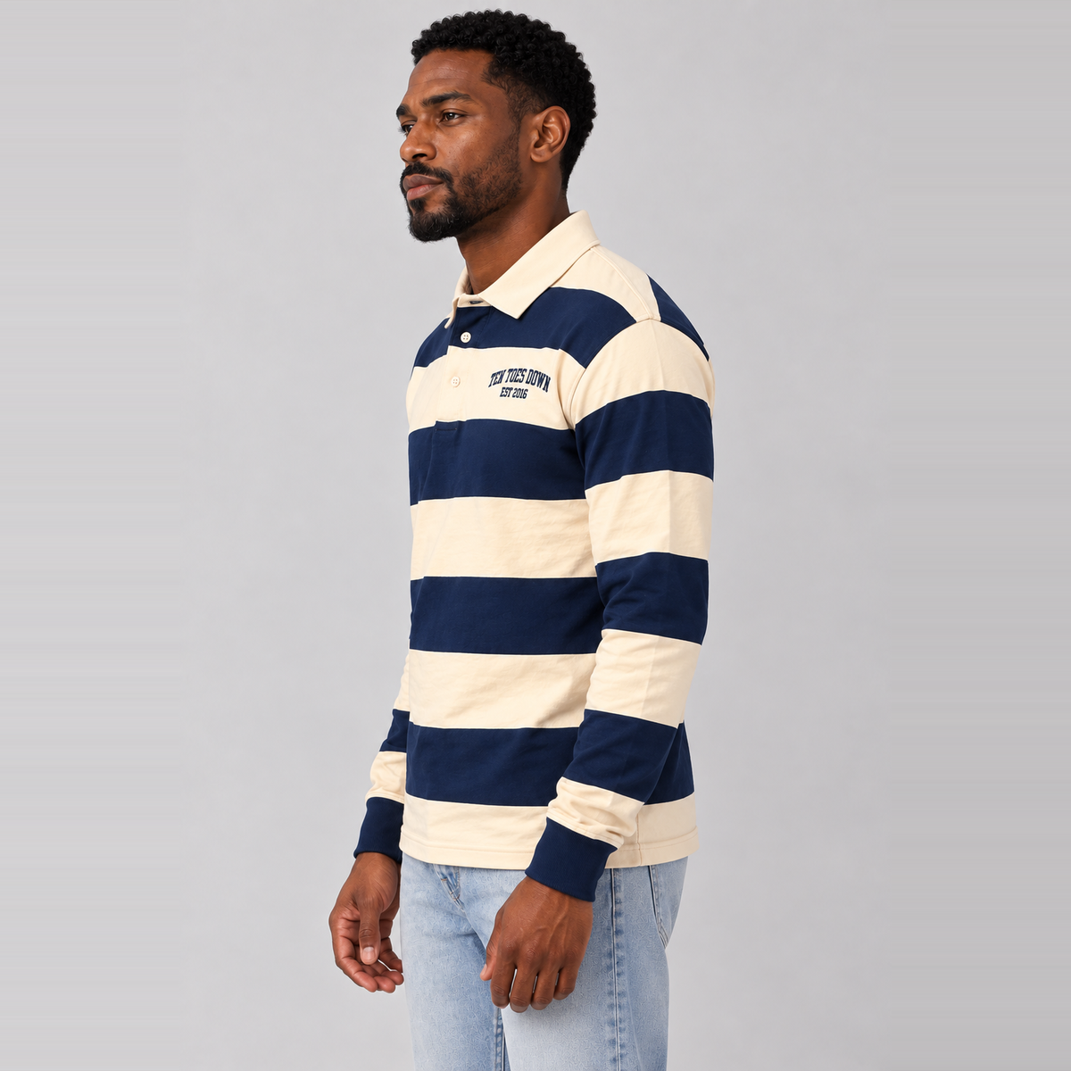A man wearing a navy and cream horizontally striped long-sleeve rugby shirt, paired with light blue jeans, standing against a solid gray studio background.