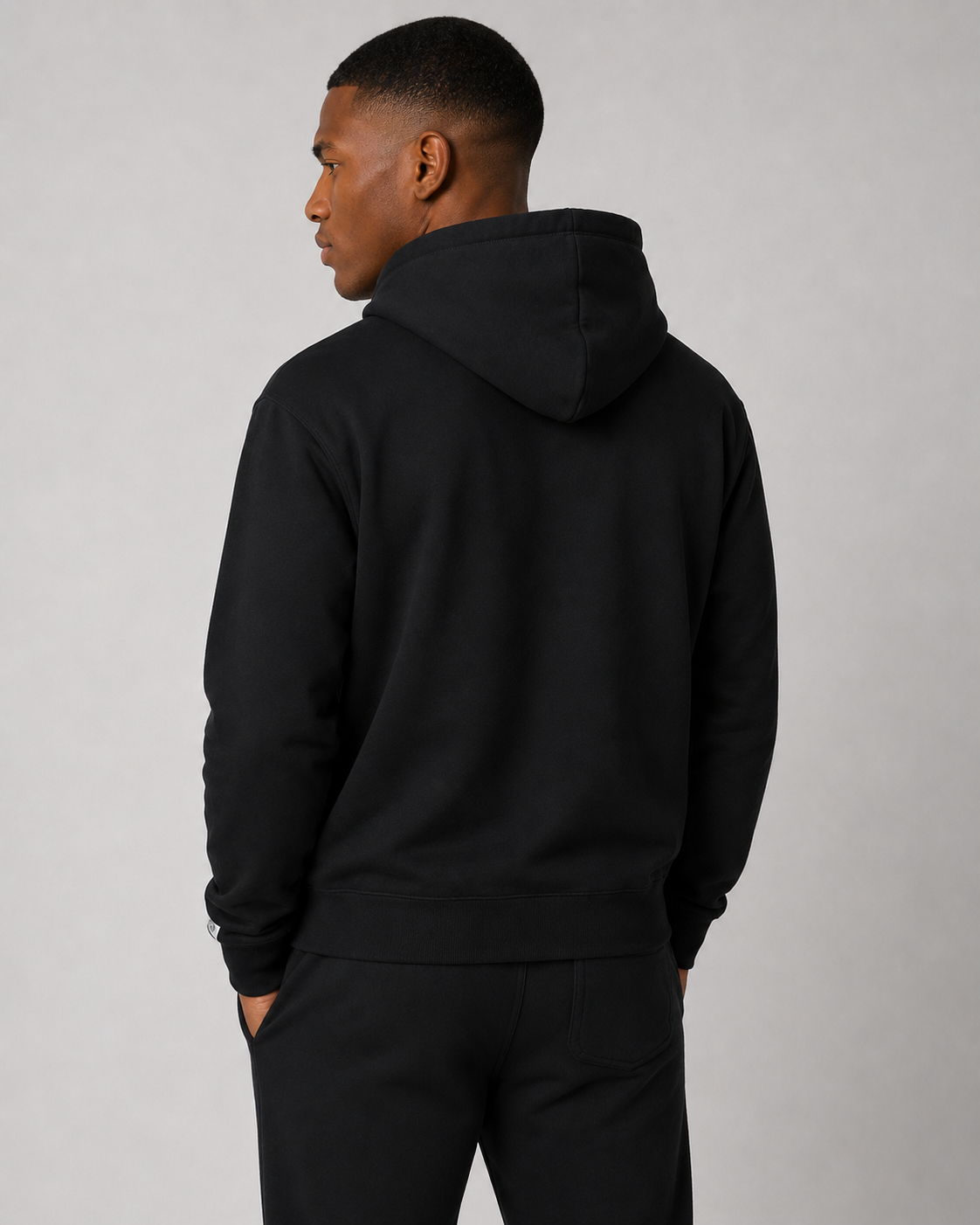 A man wearing a black pullover hoodie and matching black sweatpants, shown from the back against a plain light gray studio background.