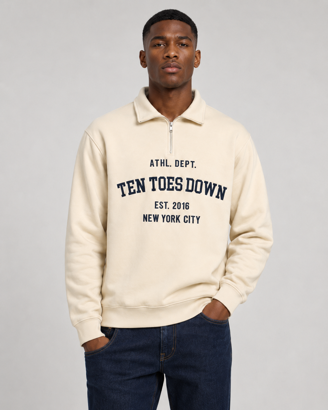 A man wearing a cream-colored quarter-zip sweatshirt with oversized 'TEN TOES DOWN' printed in navy blue on the front, paired with dark blue jeans, against a light gray studio background.