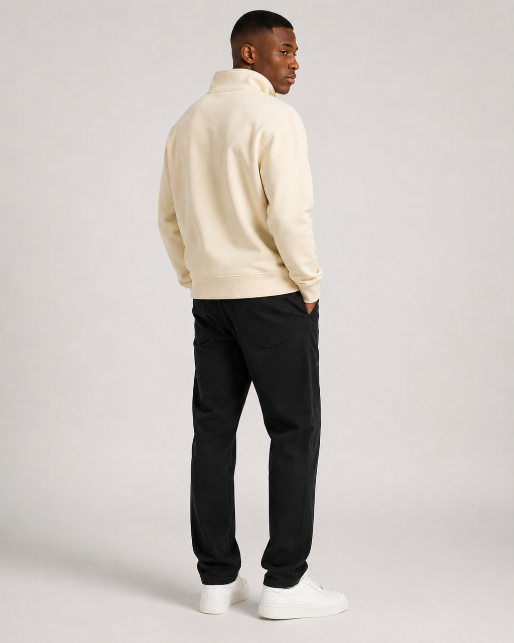 A man wearing a cream-colored quarter-zip fleece pullover, black trousers, and white sneakers, shown from a rear three-quarter view against a light gray studio background.