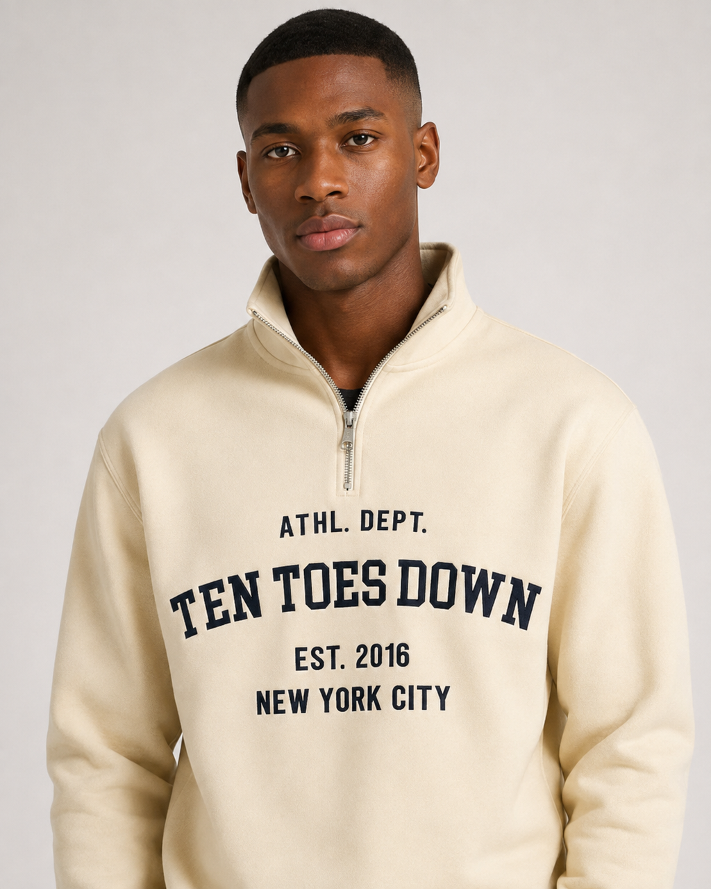 A man wearing a cream-colored quarter-zip sweatshirt with oversized  'TEN TOES DOWN' printed in navy blue collegiate lettering on the chest, set against a plain light gray background.