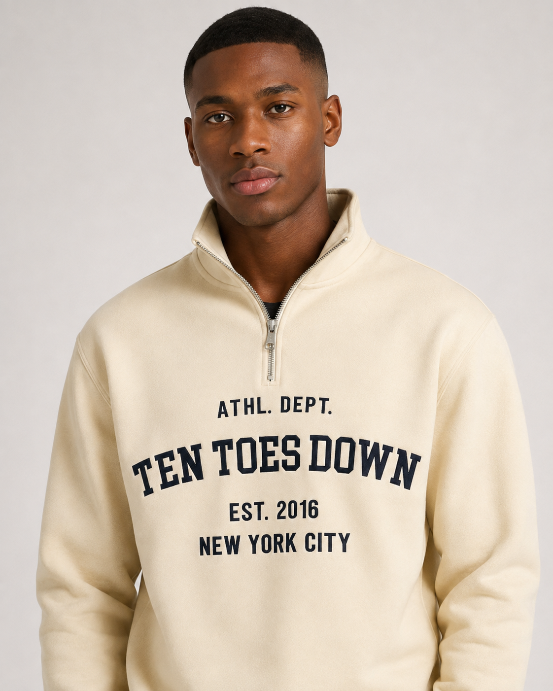 A man wearing a cream-colored quarter-zip sweatshirt with oversized  'TEN TOES DOWN' printed in navy blue collegiate lettering on the chest, set against a plain light gray background.