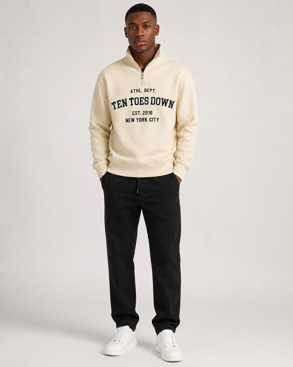 A man modeling a cream-colored quarter-zip hoodie with oversized navy text reading 'TEN TOES DOWN EST. 2016 NEW YORK CITY' on the chest, paired with black sweatpants and white sneakers, against a light gray studio background.