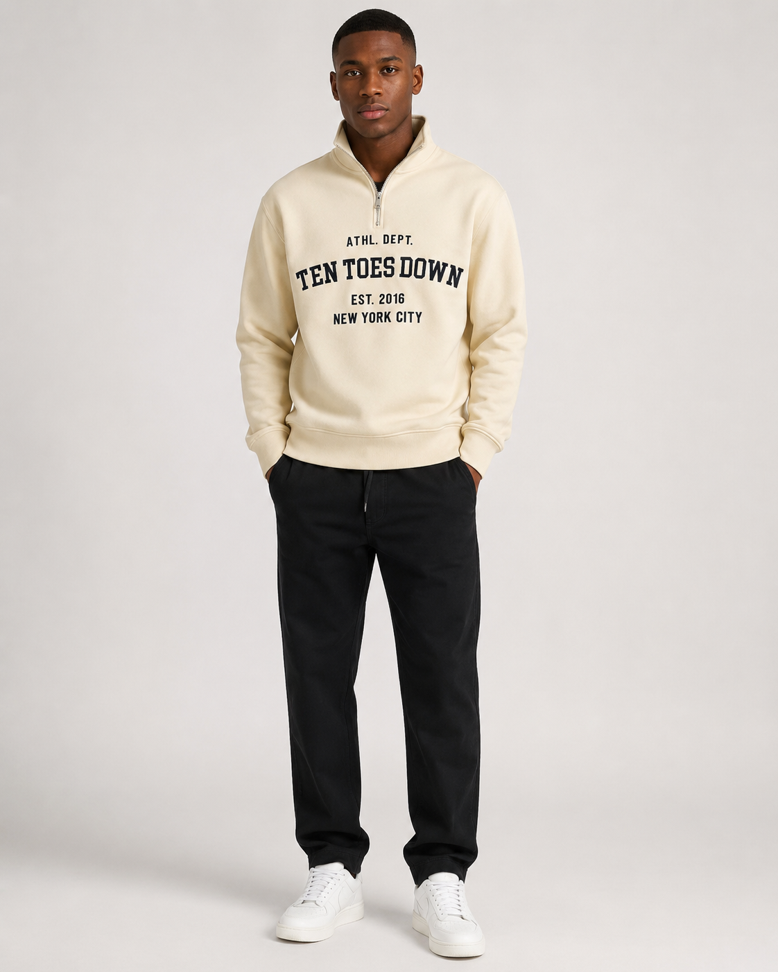 A man modeling a cream-colored quarter-zip hoodie with oversized navy text reading 'TEN TOES DOWN EST. 2016 NEW YORK CITY' on the chest, paired with black sweatpants and white sneakers, against a light gray studio background.