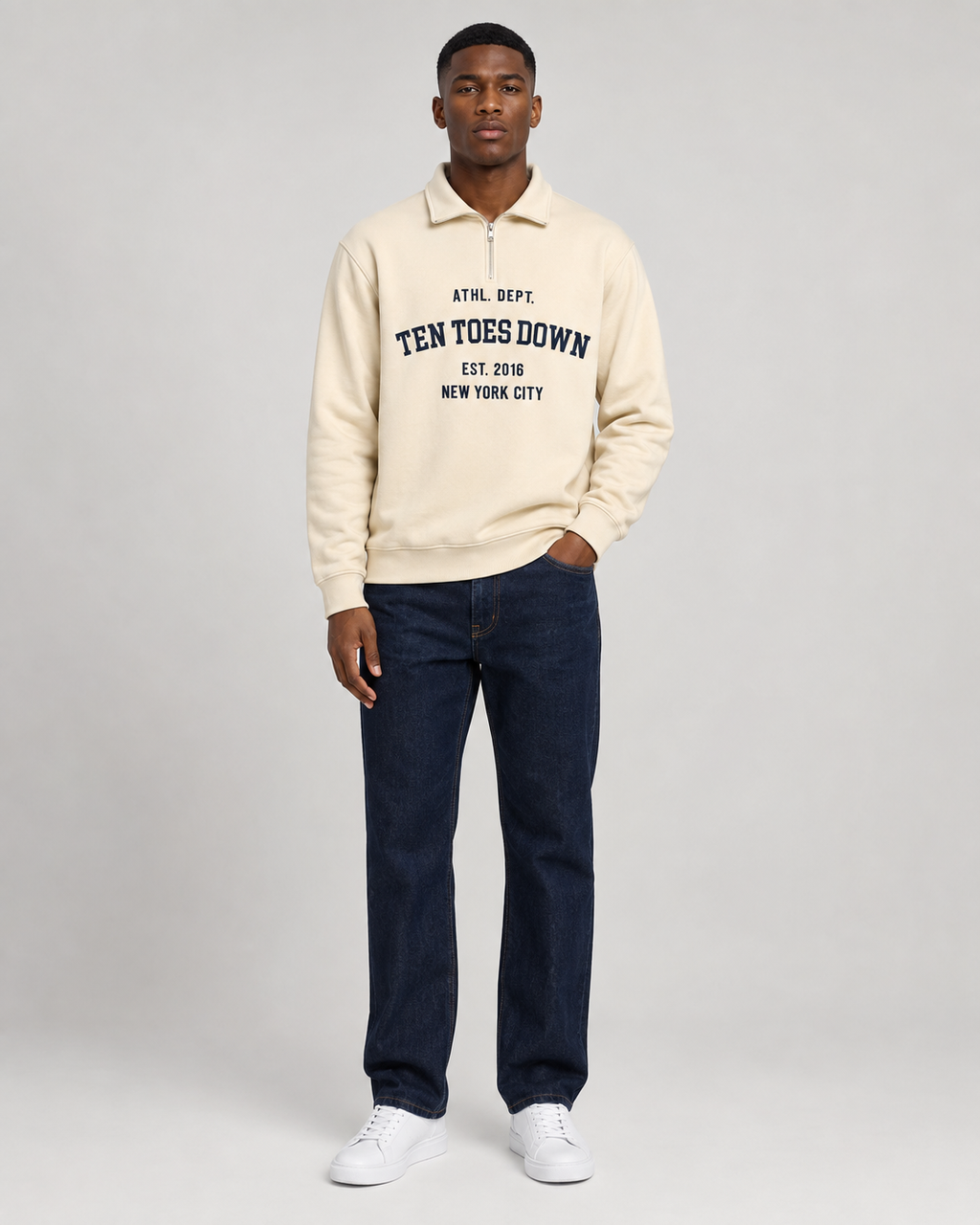 A man modeling a cream-colored quarter-zip pullover sweatshirt with oversized 'TEN TOES DOWN EST. 2016 NEW YORK CITY' printed in navy on the chest, paired with dark blue jeans and white sneakers, against a light gray studio background.
