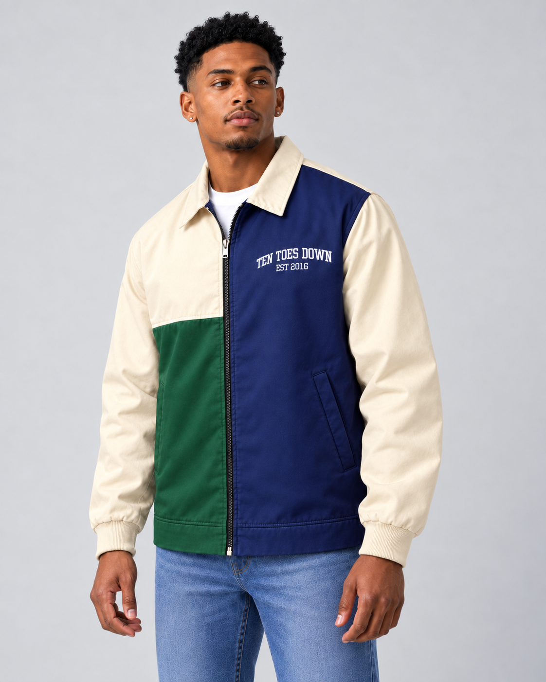 A man wearing a color-blocked varsity-style jacket with cream sleeves and collar, a green color block, and a navy blue body. The jacket features 'TEN TOES DOWN EST 2016.
