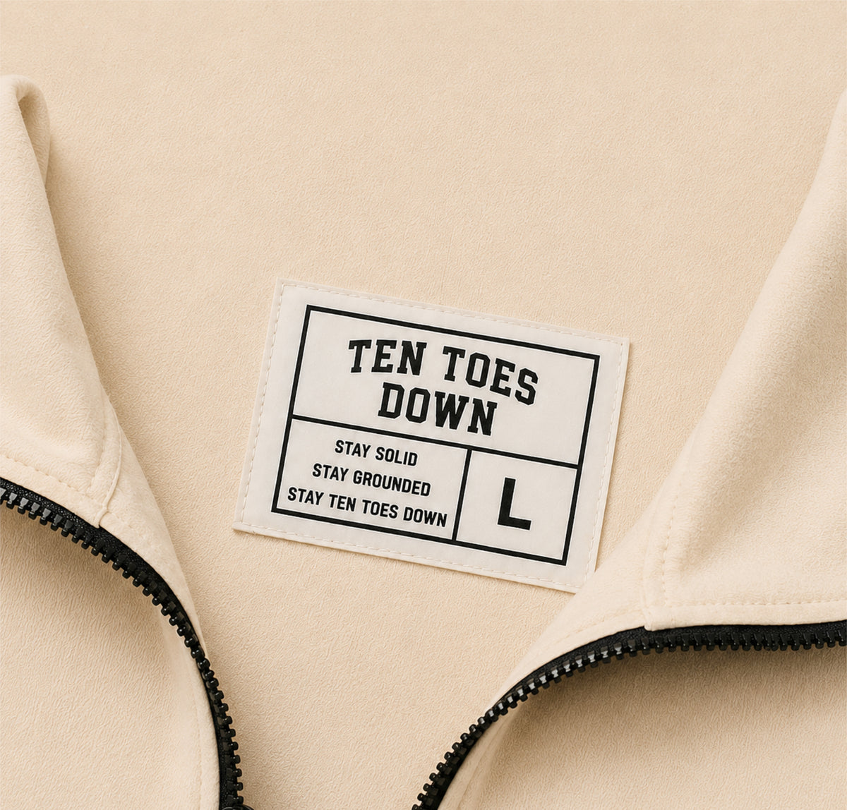Close-up of a beige zippered garment with a sewn-on white label reading 'TEN TOES DOWN' and 'STAY SOLID STAY GROUNDED STAY TEN TOES DOWN' with a large 'L'.