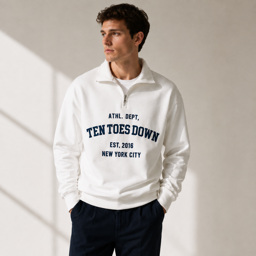 A man wearing a white quarter-zip sweatshirt with navy blue collegiate-style oversized text reading 'TEN TOES DOWN'