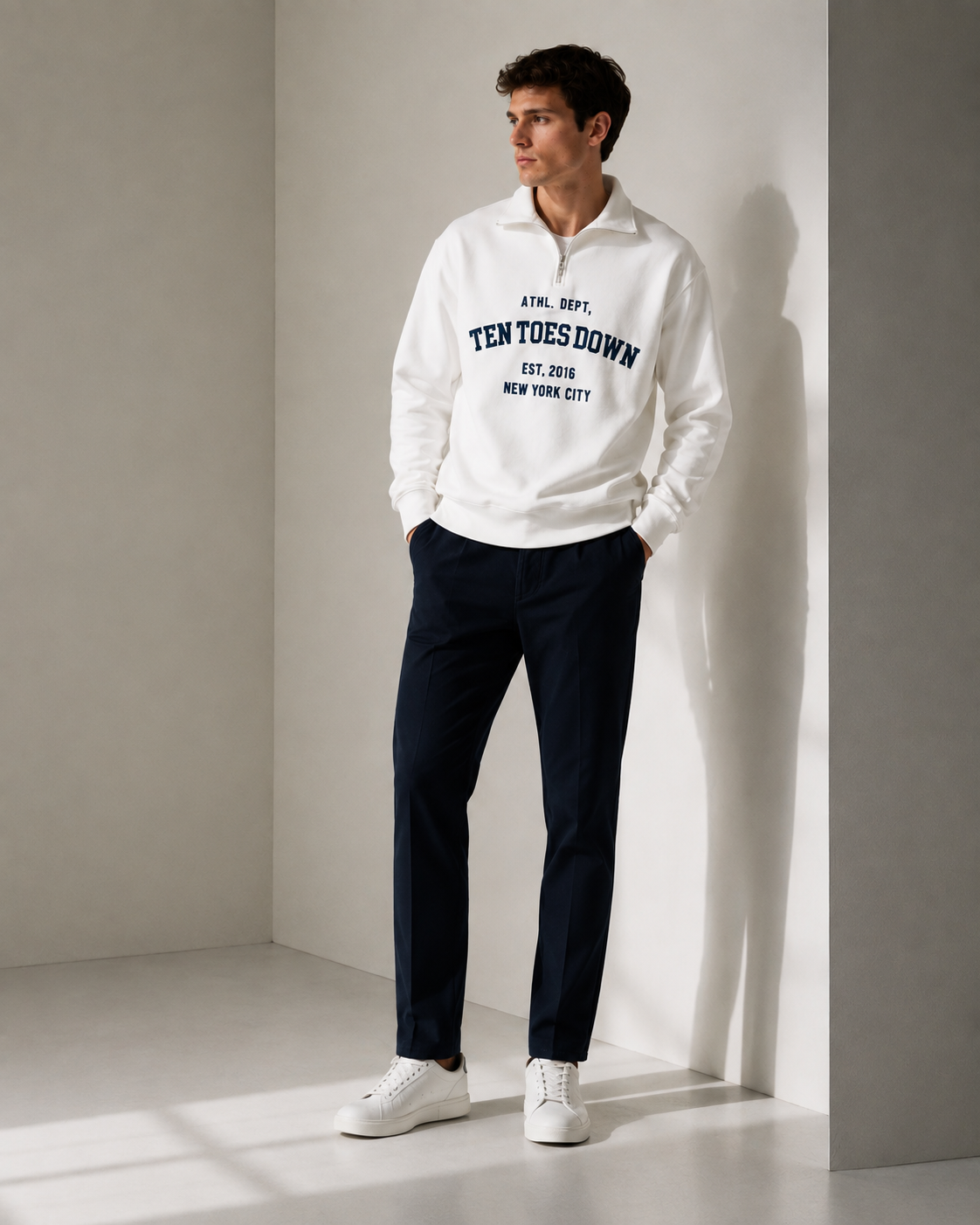 A man wearing a white quarter-zip sweatshirt with navy blue collegiate-style oversized text reading 'TEN TOES DOWN' and dark navy pants, standing in a minimalist gray corner studio setting.