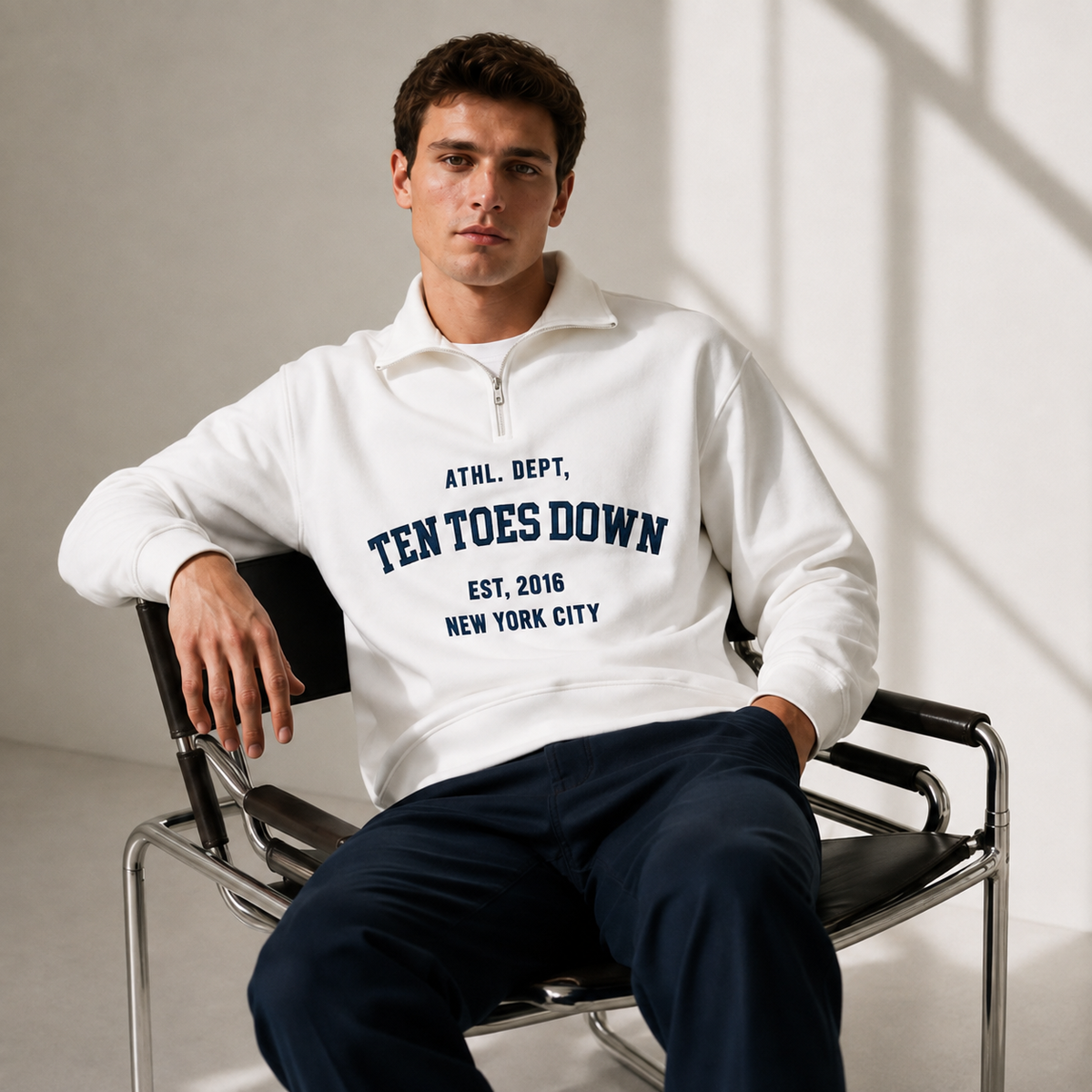 A man sits on a black chair wearing a white quarter-zip sweatshirt with navy blue oversized text reading 'TEN TOES DOWN'