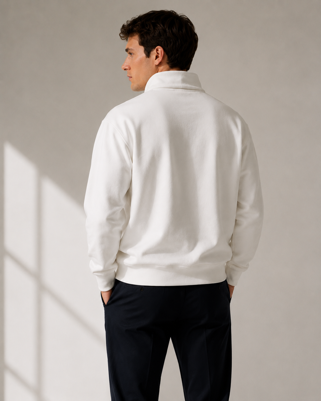 A man wearing a white quarter zip sweatshirt and dark navy pants, shown from the back against a neutral light gray wall with soft window shadow patterns.